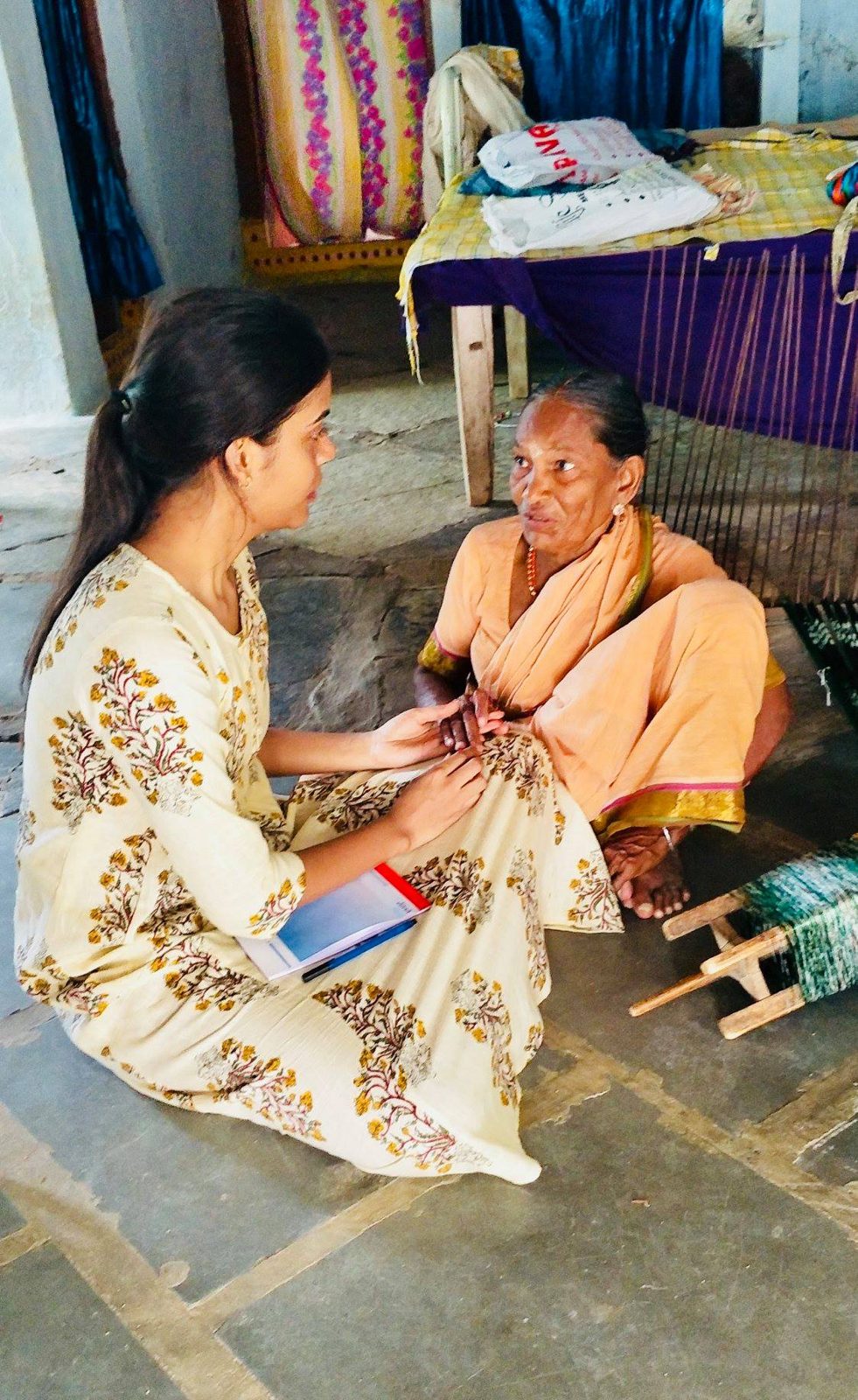 The founder sitting on the floor of a weaver’s home, holding the hands of an elderly woman as she talks