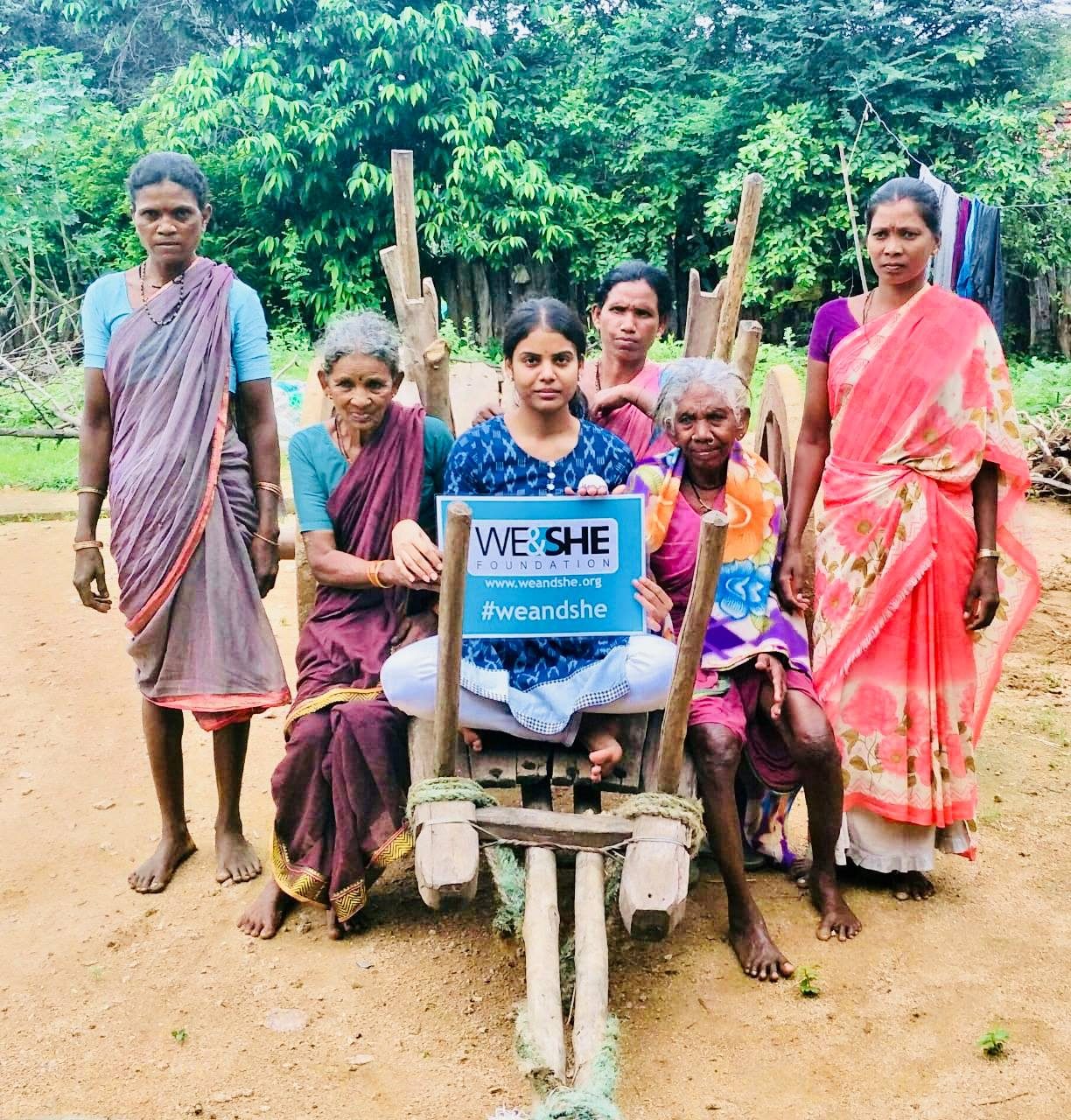 Elderly village women gathered around the founder, who holds a We & She sign